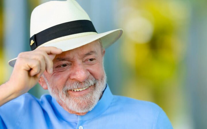 O presidente Lula, de chapéu panamá e camisa azul, sorri e segura a aba do chapéu