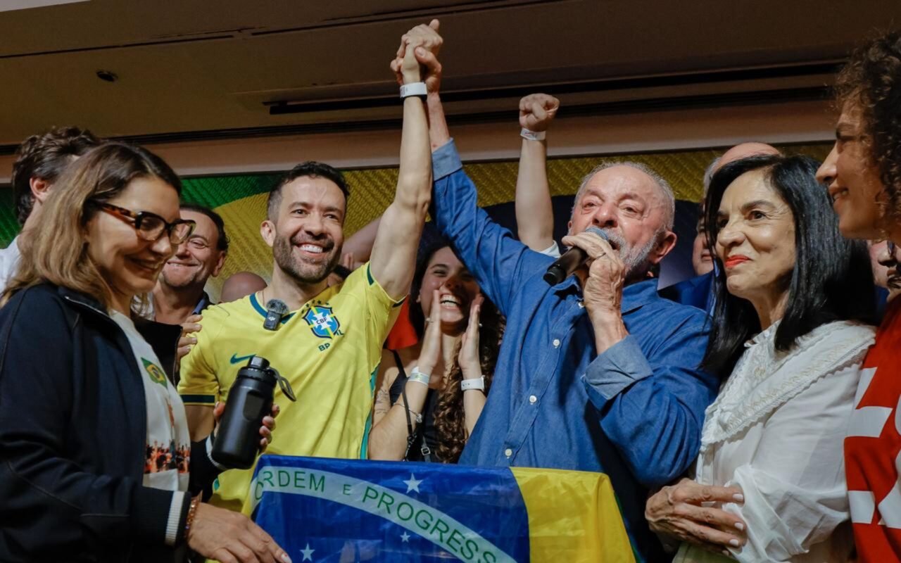 O presidente Lula, ao microfone e rodeado de apoiadores, com a bandeira do Brasil à frente