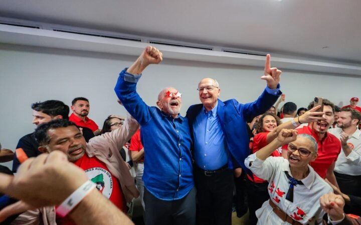 O presidente Lula, de braços erguidos, comemora ao lado de aliados em sala cheia