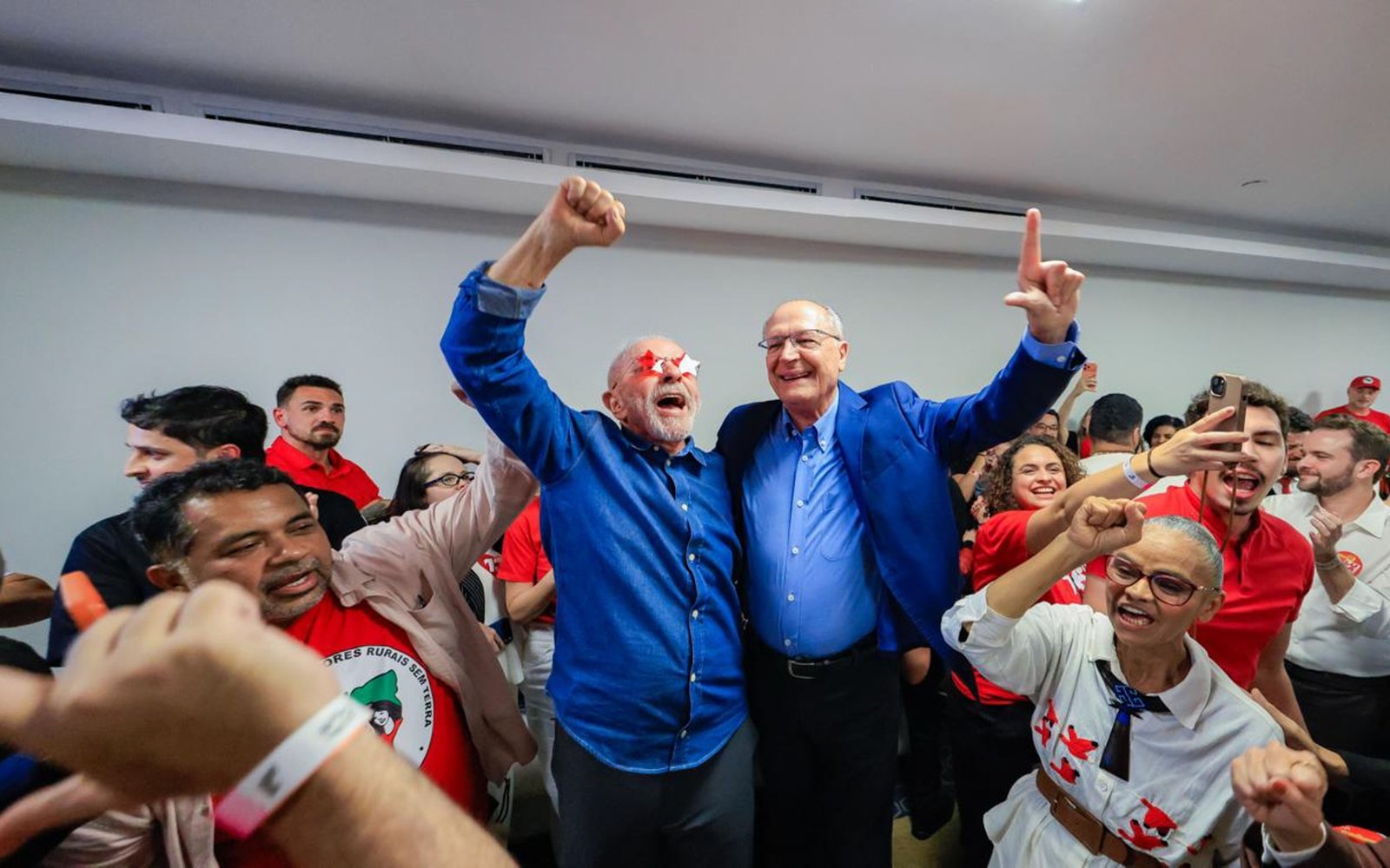 O presidente Lula, de braços erguidos, comemora ao lado de aliados em sala cheia