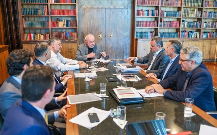 Lula à cabeceira de uma mesa de reunião com ministros, em sala com estantes de livros