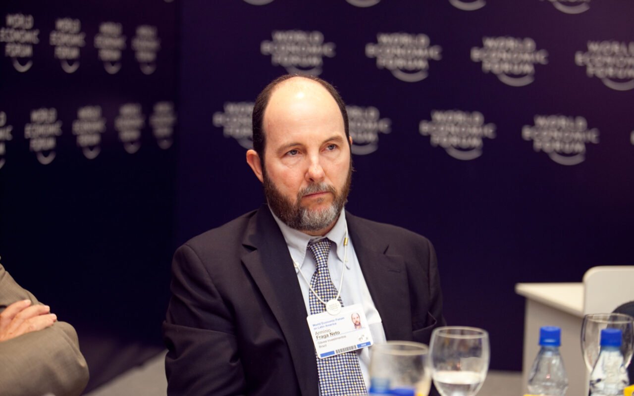 Armínio Fraga, de terno escuro e gravata, sentado diante de um painel azul-escuro do Fórum Econômico Mundial, com crachá no pescoço.