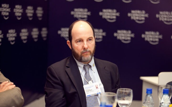 Armínio Fraga, de terno escuro e gravata, sentado diante de um painel azul-escuro do Fórum Econômico Mundial, com crachá no pescoço.