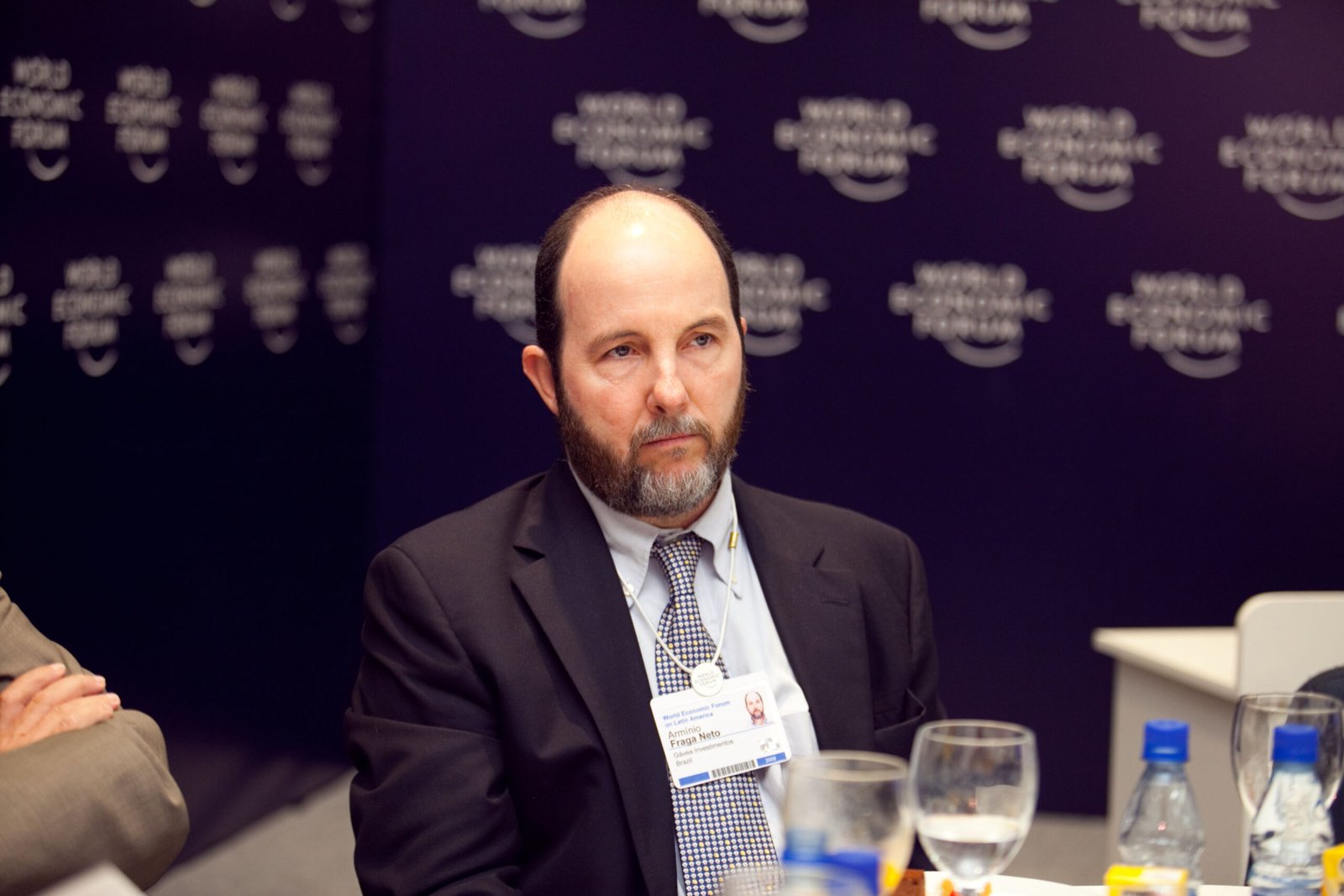 Armínio Fraga, de terno escuro e gravata, sentado diante de um painel azul-escuro do Fórum Econômico Mundial, com crachá no pescoço.