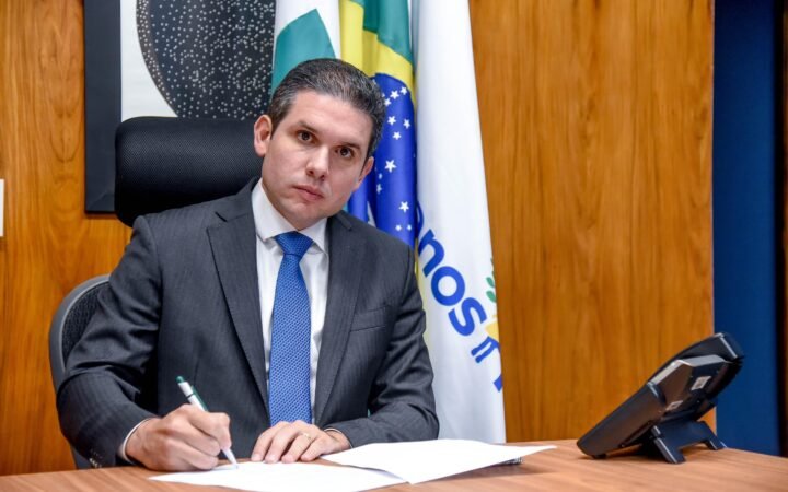 Hugo Motta, de terno cinza e gravata azul, assina um documento sentado a uma mesa, com bandeira do Brasil ao fundo.