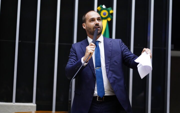 O então deputado Eduardo Bolsonaro, de terno azul e gravata azul, discursa ao microfone no plenário da Câmara com papéis na mão.