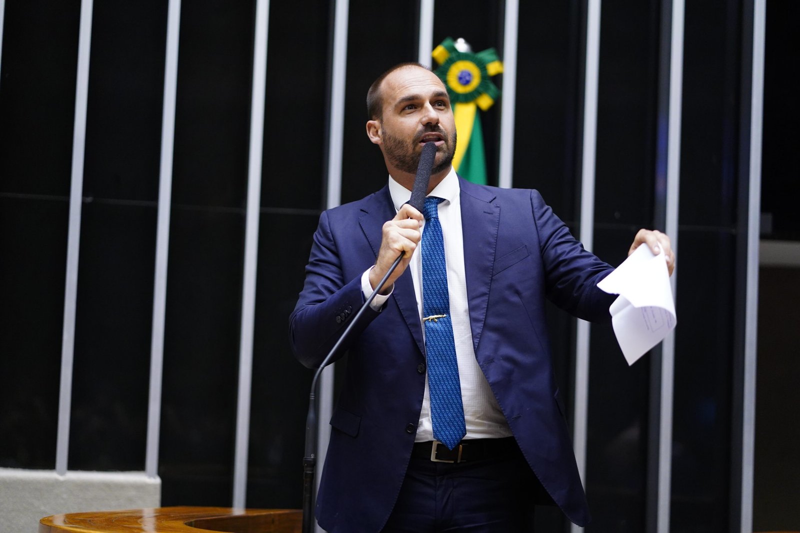 O então deputado Eduardo Bolsonaro, de terno azul e gravata azul, discursa ao microfone no plenário da Câmara com papéis na mão.
