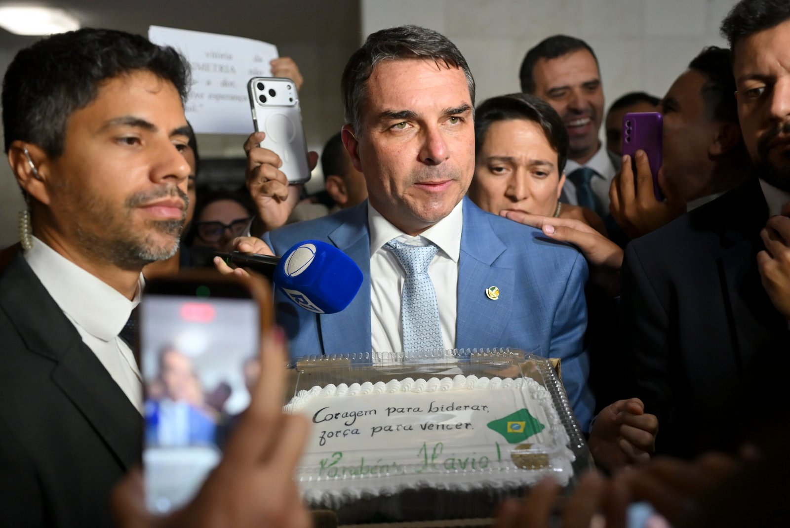 Senador Flávio Bolsonaro, de terno azul, fala a repórteres cercado por microfones e celulares, com um bolo à frente.