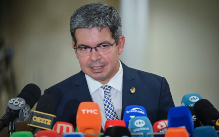 O senador Randolfe Rodrigues, de óculos e terno azul, fala diante de vários microfones de emissoras de TV.