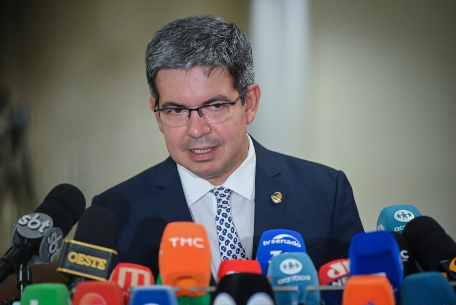 O senador Randolfe Rodrigues, de óculos e terno azul, fala diante de vários microfones de emissoras de TV.