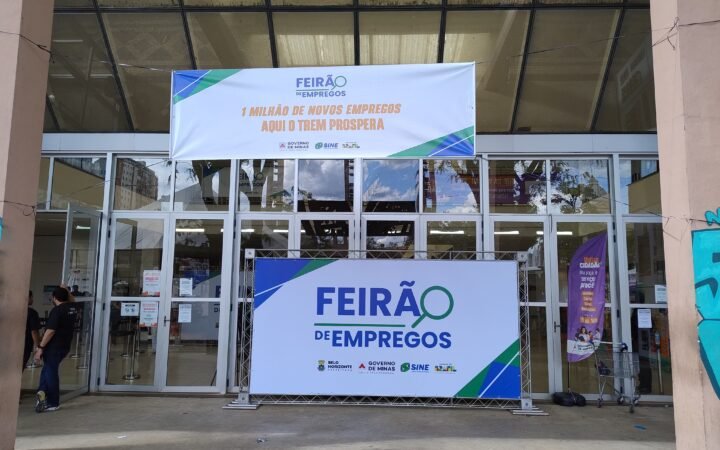 Entrada de um feirão de empregos em Belo Horizonte, com faixa e painel do evento diante das portas de vidro de um centro cultural; um funcionário aparece à esquerda.