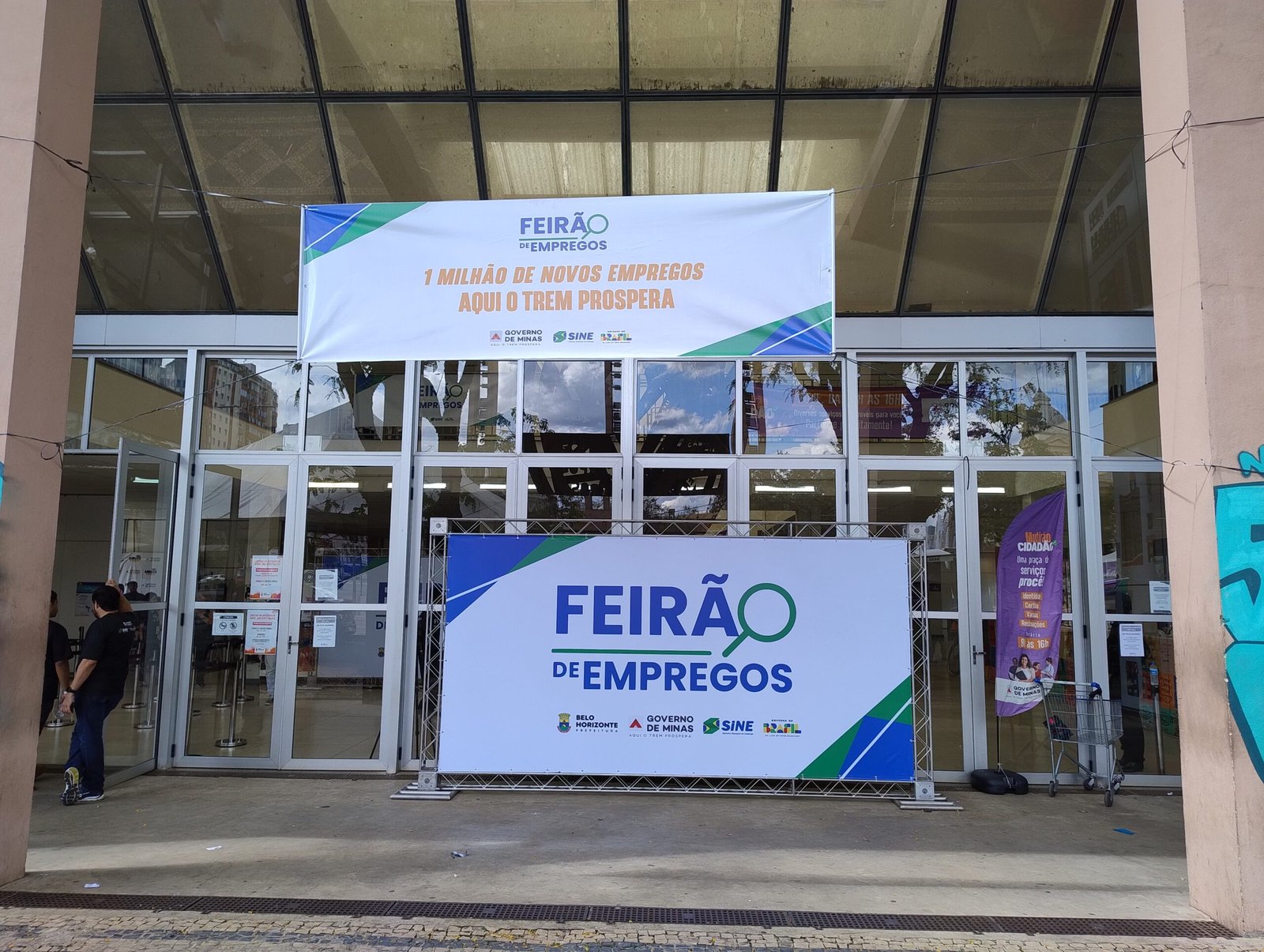 Entrada de um feirão de empregos em Belo Horizonte, com faixa e painel do evento diante das portas de vidro de um centro cultural; um funcionário aparece à esquerda.