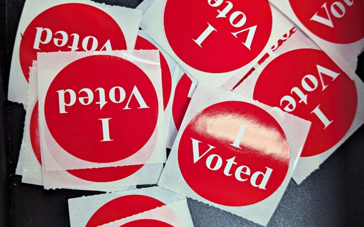 Pilha de adesivos vermelhos com a inscrição 'I Voted' sobre uma superfície escura.