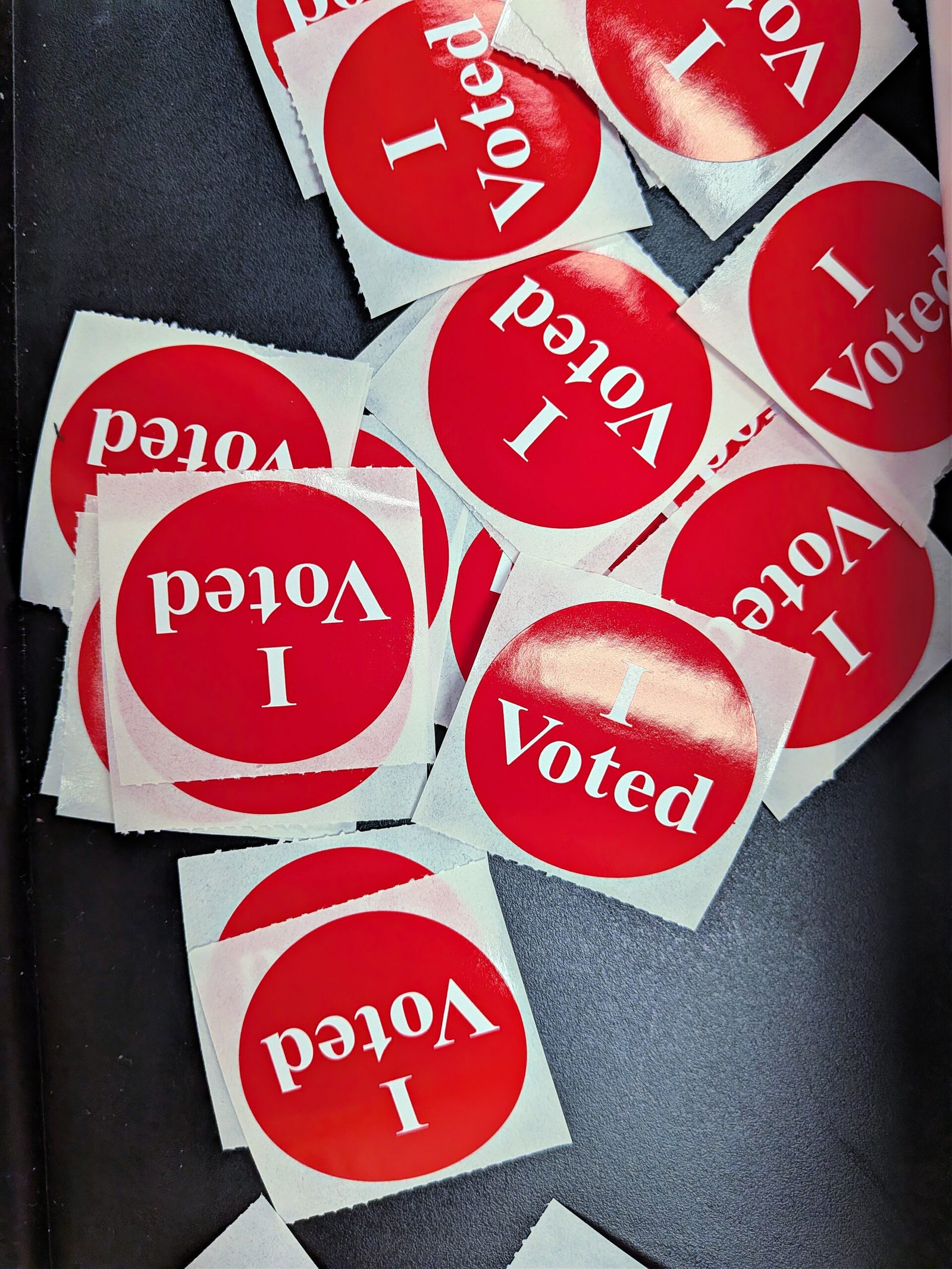 Pilha de adesivos vermelhos com a inscrição 'I Voted' sobre uma superfície escura.