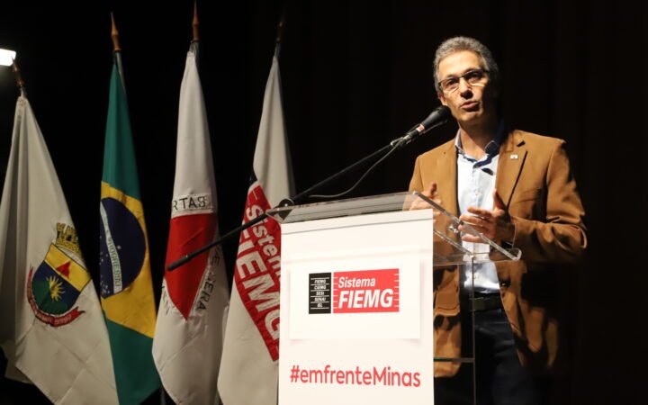 Romeu Zema, de paletó marrom, fala ao microfone em um púlpito, ao lado de bandeiras do Brasil e de Minas Gerais