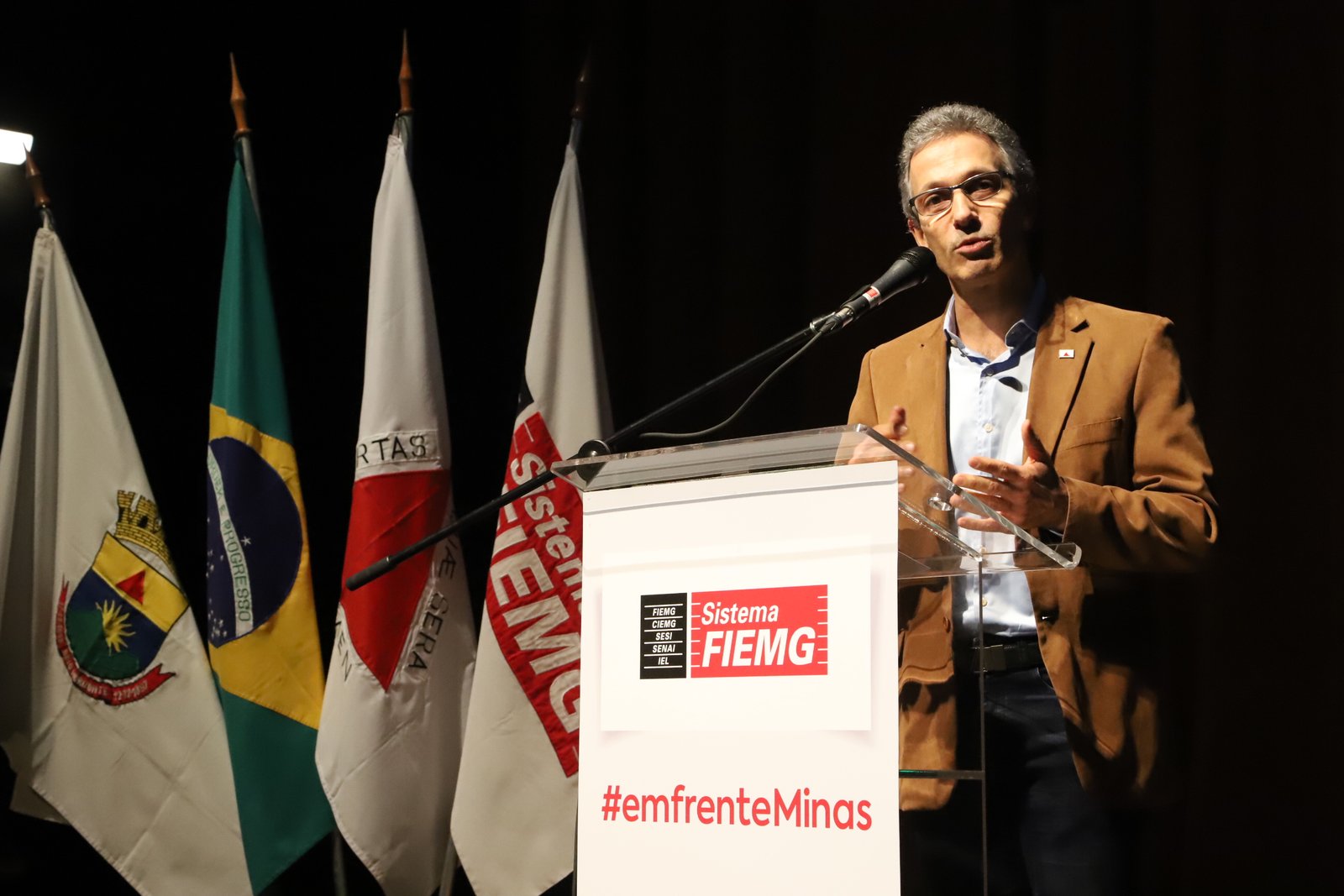 Romeu Zema, de paletó marrom, fala ao microfone em um púlpito, ao lado de bandeiras do Brasil e de Minas Gerais