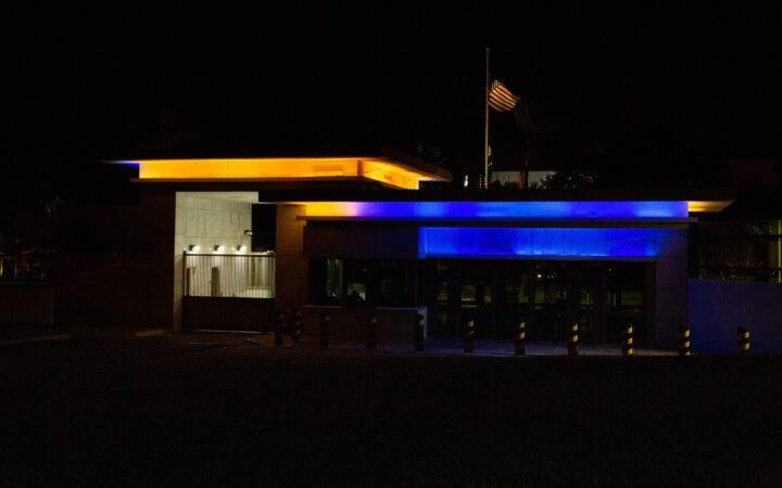 Entrada da embaixada dos Estados Unidos em Brasília à noite, iluminada em azul e amarelo, com a bandeira americana ao fundo