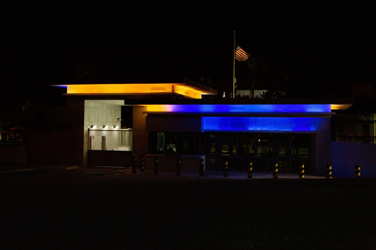 Entrada da embaixada dos Estados Unidos em Brasília à noite, iluminada em azul e amarelo, com a bandeira americana ao fundo