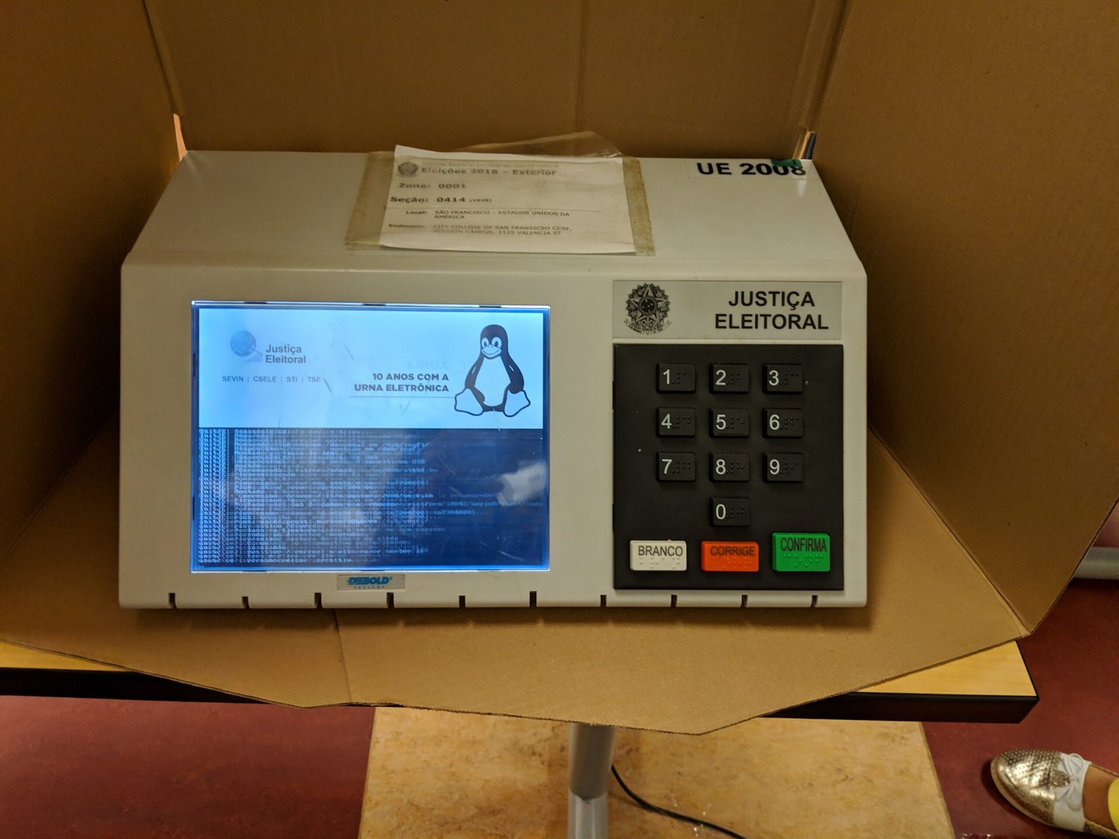Urna eletrônica brasileira com tela e teclado numérico dentro de cabine de papelão