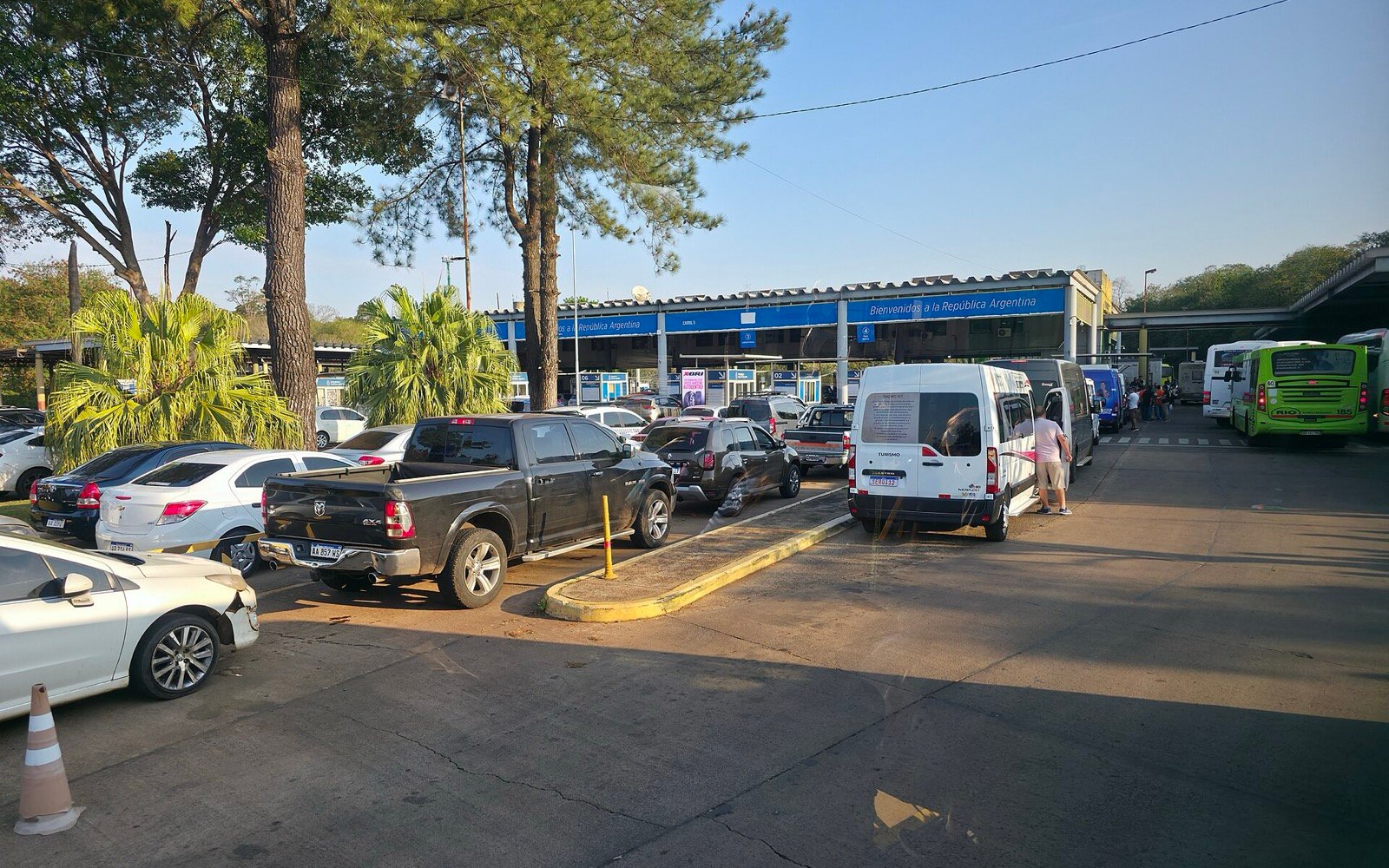 Pátio do posto de controle argentino em Puerto Iguazú, com carros, uma van e ônibus e a faixa azul "Bienvenidos a la República Argentina" sobre o prédio da aduana.