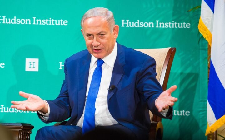 Benjamin Netanyahu, de terno azul e gravata azul, gesticula sentado diante de painel verde