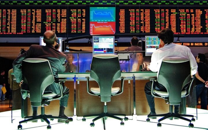 Dois operadores sentados de costas diante do painel de cotações da Bolsa de Valores de São Paulo, com gráficos em queda na tela central