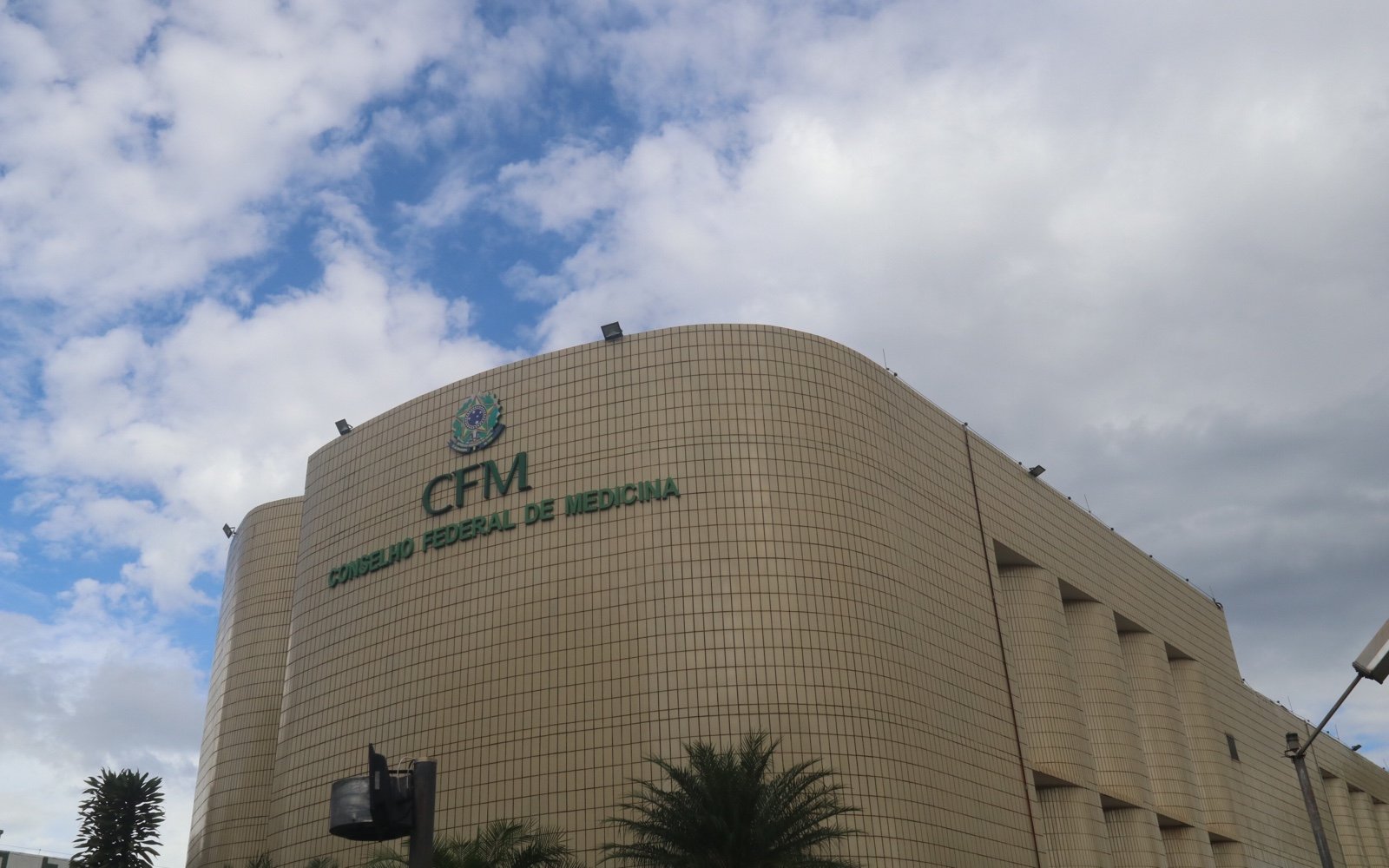 Fachada curva revestida de pastilhas da sede do Conselho Federal de Medicina, em Brasília, com o nome e o brasão da entidade e céu com nuvens.