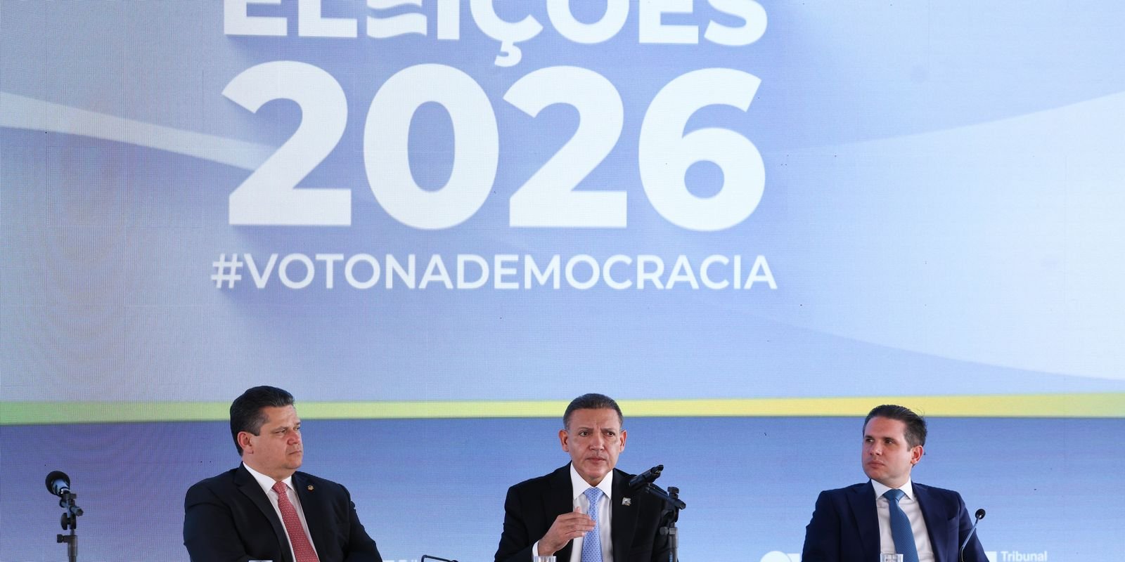 Davi Alcolumbre, o ministro Nunes Marques e Hugo Motta sentados à mesa diante de um painel com a inscrição Eleições 2026, durante entrevista coletiva no TSE