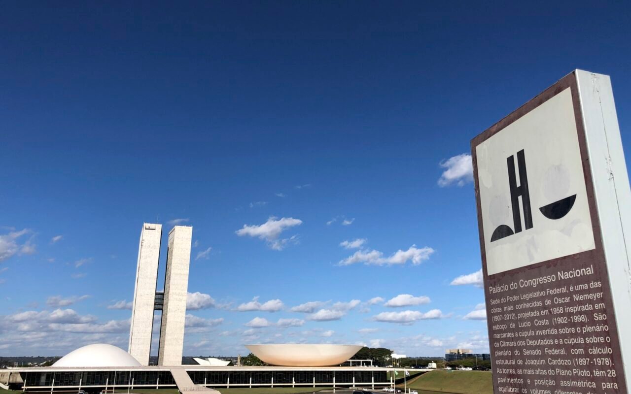 Palácio do Congresso Nacional, em Brasília, com as torres gêmeas e as cúpulas sob céu azul; à direita, placa informativa.
