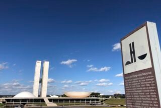 Palácio do Congresso Nacional, em Brasília, com as torres gêmeas e as cúpulas sob céu azul; à direita, placa informativa.