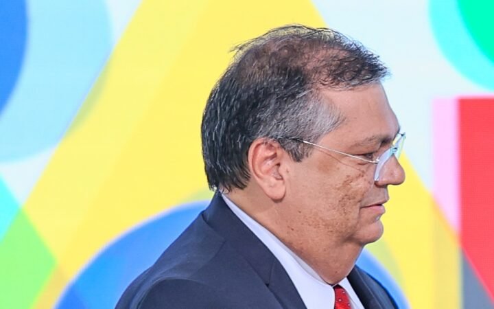 Flávio Dino, de perfil e de óculos, terno escuro e gravata vermelha, em cerimônia de 2023