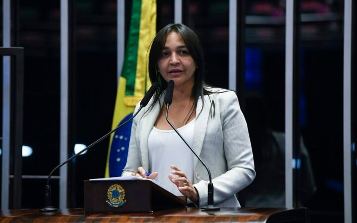 A senadora Eliziane Gama discursa ao microfone, com a bandeira do Brasil ao fundo