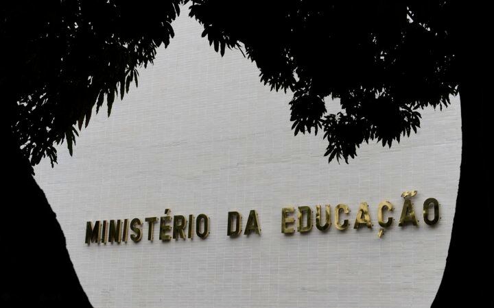 Letreiro dourado Ministério da Educação na parede de pastilhas brancas do prédio, emoldurado por galhos de árvore
