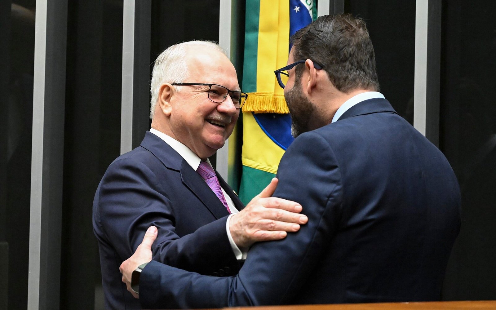 O presidente do STF, Edson Fachin, de óculos e sorrindo, cumprimenta de costas o presidente da Câmara, Hugo Motta, diante de uma bandeira do Brasil.
