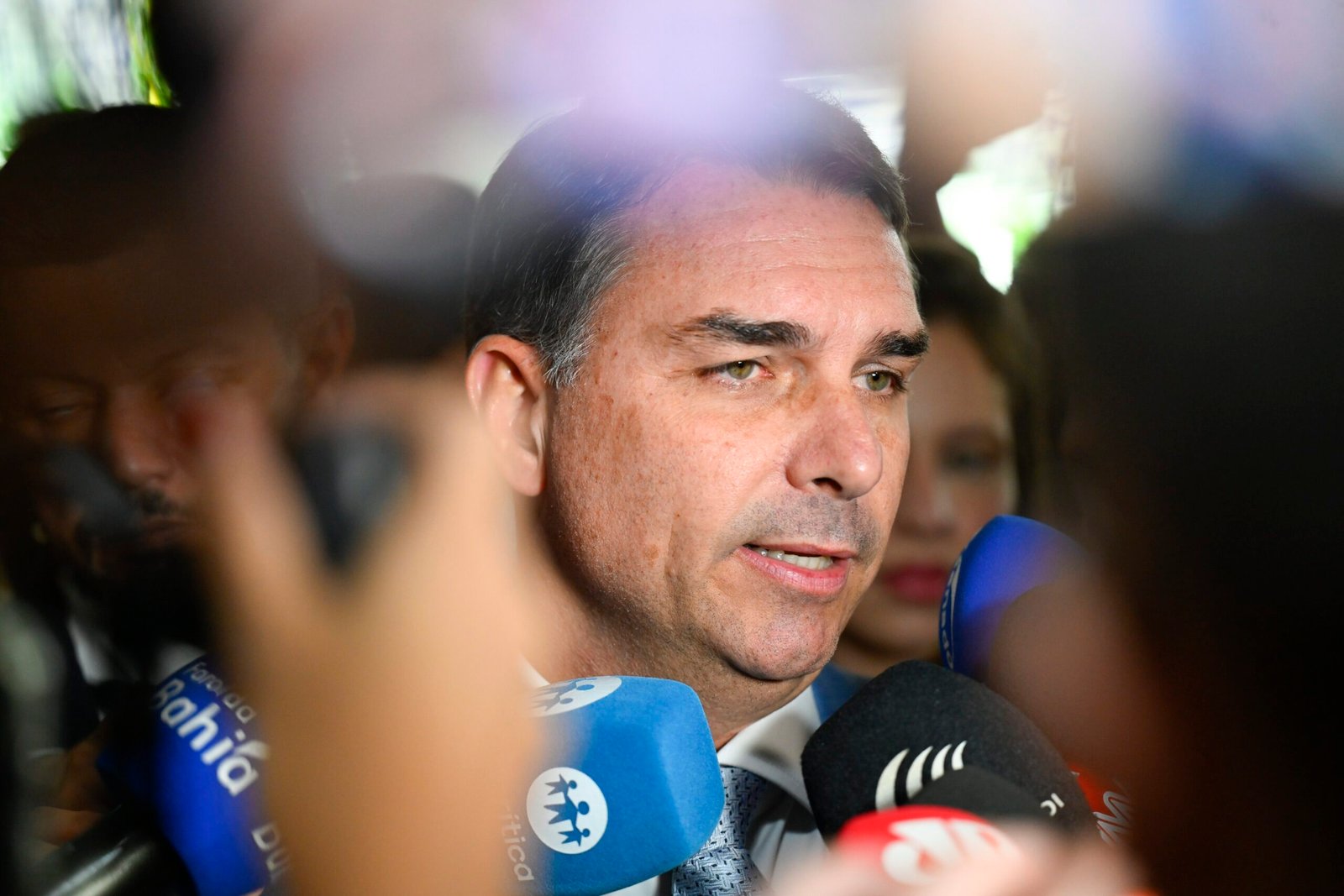 Senador Flávio Bolsonaro, em close, fala a jornalistas cercado por microfones, no Senado.