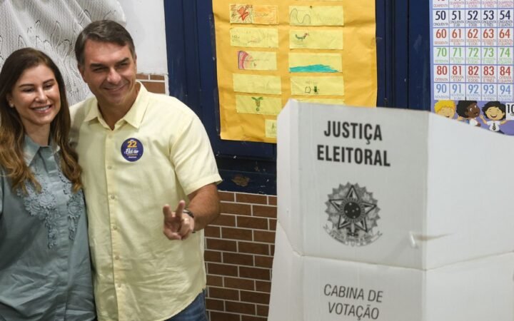 Flávio Bolsonaro, de camisa amarela, faz sinal com as mãos ao lado da mulher, Fernanda Bolsonaro, junto à cabine de votação da Justiça Eleitoral, no Rio de Janeiro