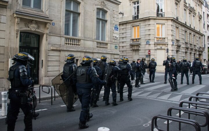 Policiais franceses de capacete e colete, com escudos, caminham por uma rua de Paris entre prédios de pedra