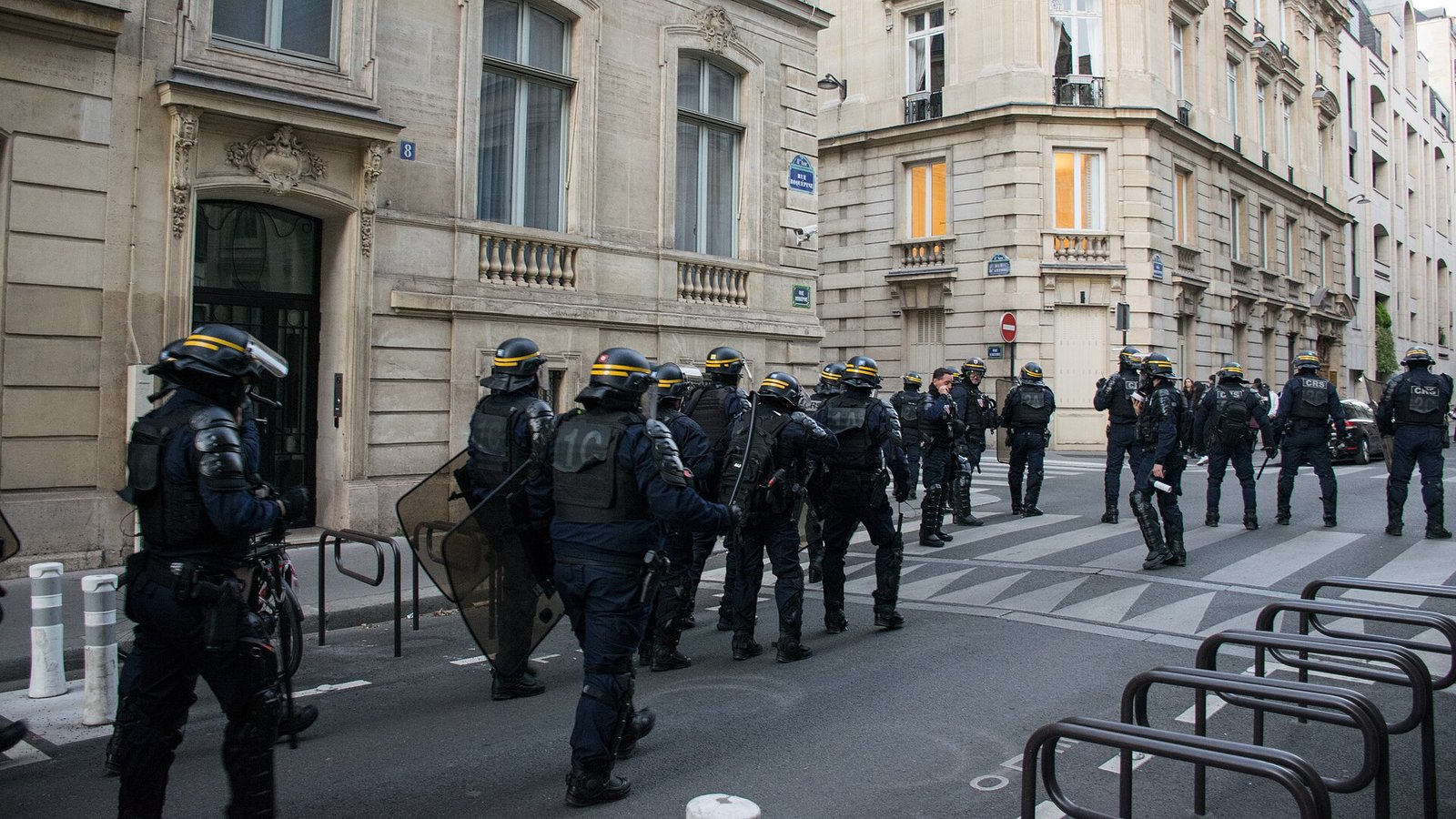 Policiais franceses de capacete e colete, com escudos, caminham por uma rua de Paris entre prédios de pedra