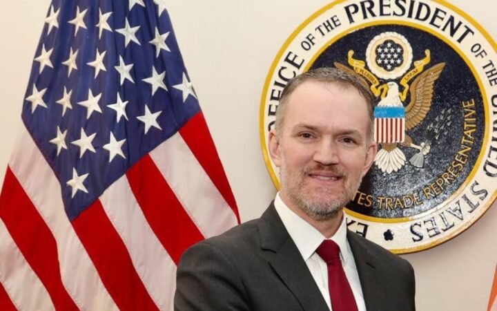 Jamieson Greer, representante comercial dos Estados Unidos, de terno escuro e gravata vermelha, diante da bandeira americana e do selo do Escritório do Representante Comercial