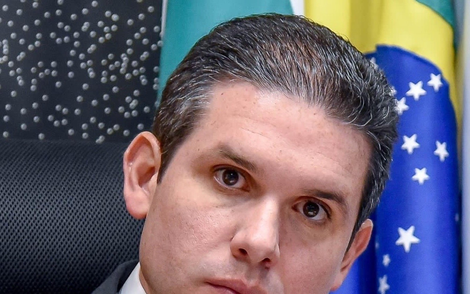 Retrato do deputado Hugo Motta, de terno cinza e gravata azul, diante de uma bandeira do Brasil