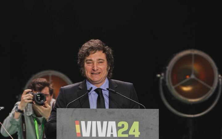 Javier Milei, de terno escuro, sorri atrás de um púlpito com microfones