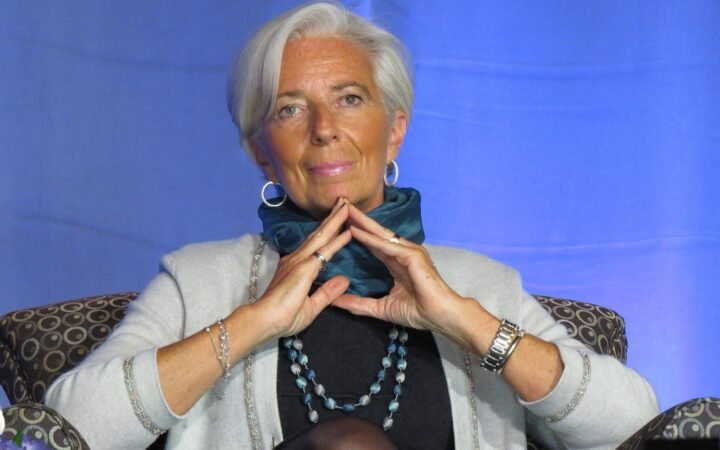 Christine Lagarde sentada, com as mãos unidas diante do queixo, diante de um fundo azul
