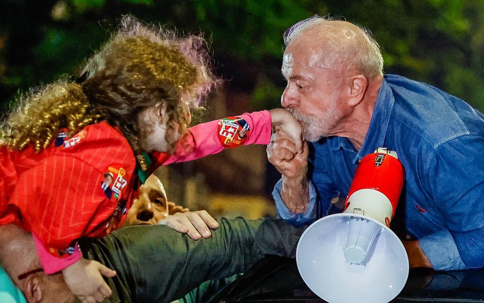 O presidente Lula, de camisa jeans e com um megafone, beija a mão de uma criança em ato de comemoração