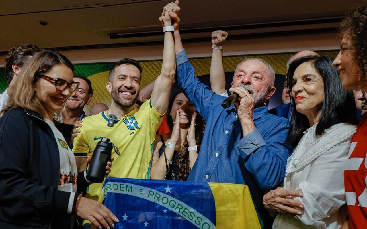 Lula, de camisa azul e microfone na mão, tem o braço erguido por apoiadores durante o discurso da reeleição, ao lado de uma bandeira do Brasil