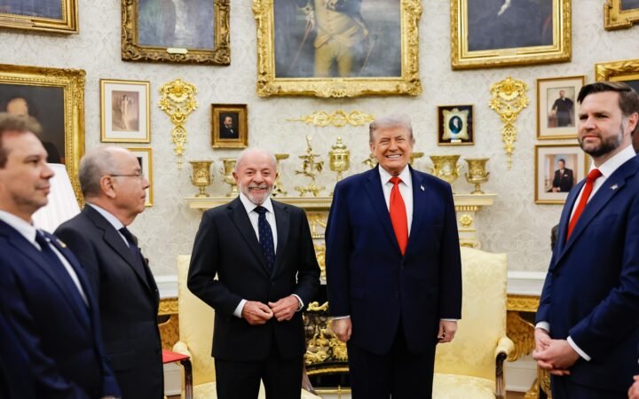 Lula e Donald Trump lado a lado, sorrindo, no Salão Oval da Casa Branca, com o vice J.D. Vance e outros presentes ao redor.