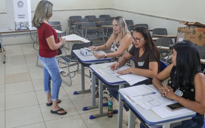 Três mesárias sentadas a mesas escolares conferem documentos de eleitores em uma sala de aula usada como seção eleitoral, com a cabine de votação ao fundo, em Brasília