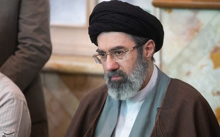 Retrato de Mojtaba Khamenei, de turbante preto, barba grisalha e óculos, sentado em um salão