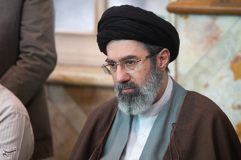 Retrato de Mojtaba Khamenei, de turbante preto, barba grisalha e óculos, sentado em um salão
