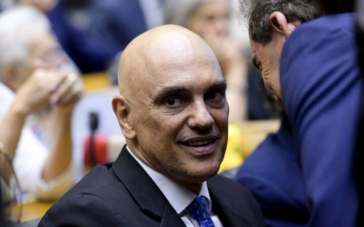O ministro do STF Alexandre de Moraes, de terno escuro e gravata azul, sorri sentado no plenário da Câmara dos Deputados
