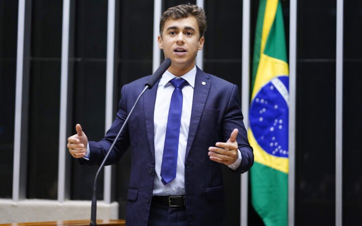 Deputado Nikolas Ferreira discursando na tribuna do plenário da Câmara, ao lado da bandeira do Brasil